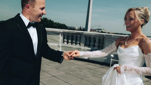 The groom reaches for the bride's hand on the rooftop terrace, her lace sleeves and pearls catching the midday sun