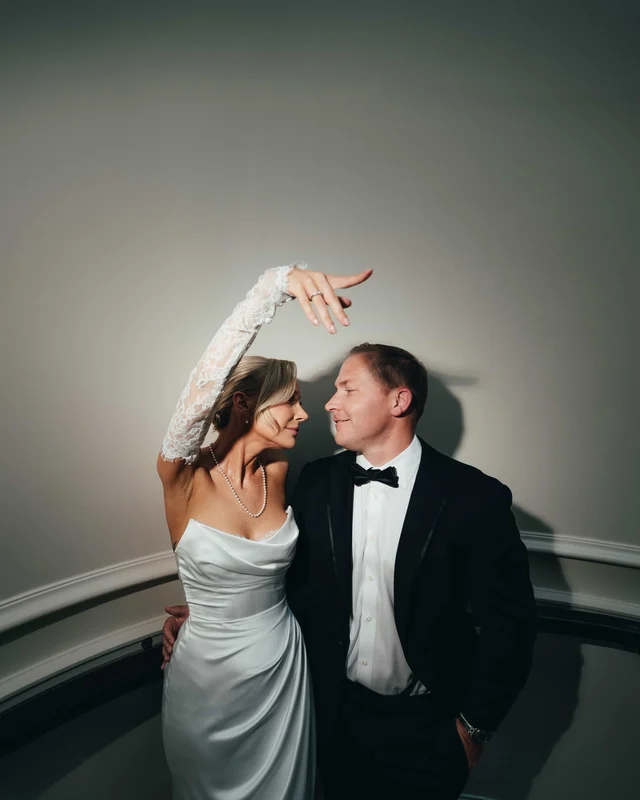 The bride arcs a lace-gloved arm overhead while the groom leans in, a single spotlight throwing their shadow on the curved wall