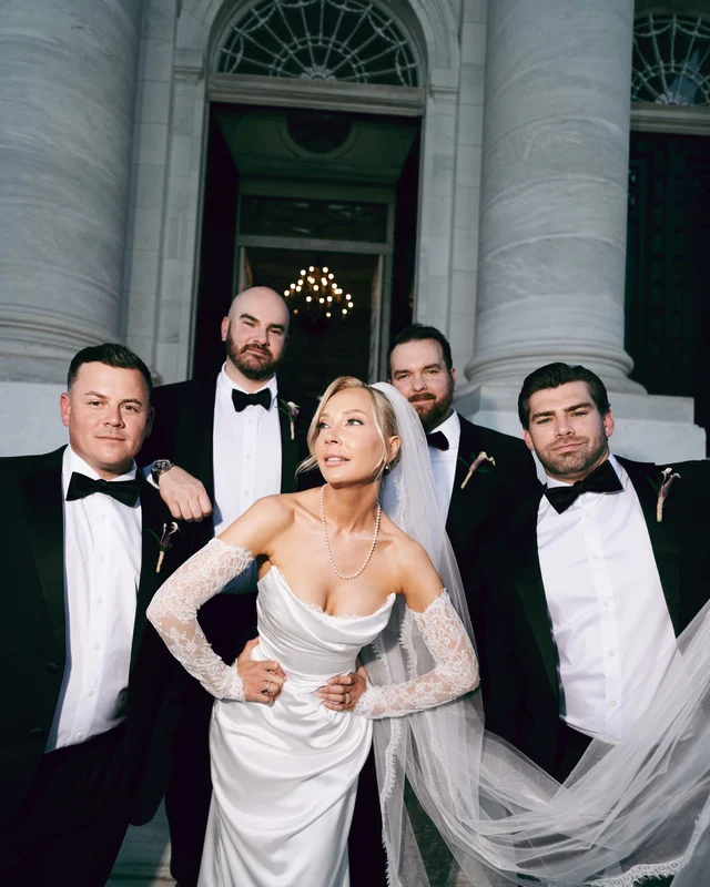 The bride stands hands on hips among four groomsmen between colossal marble columns, her veil sweeping across the steps