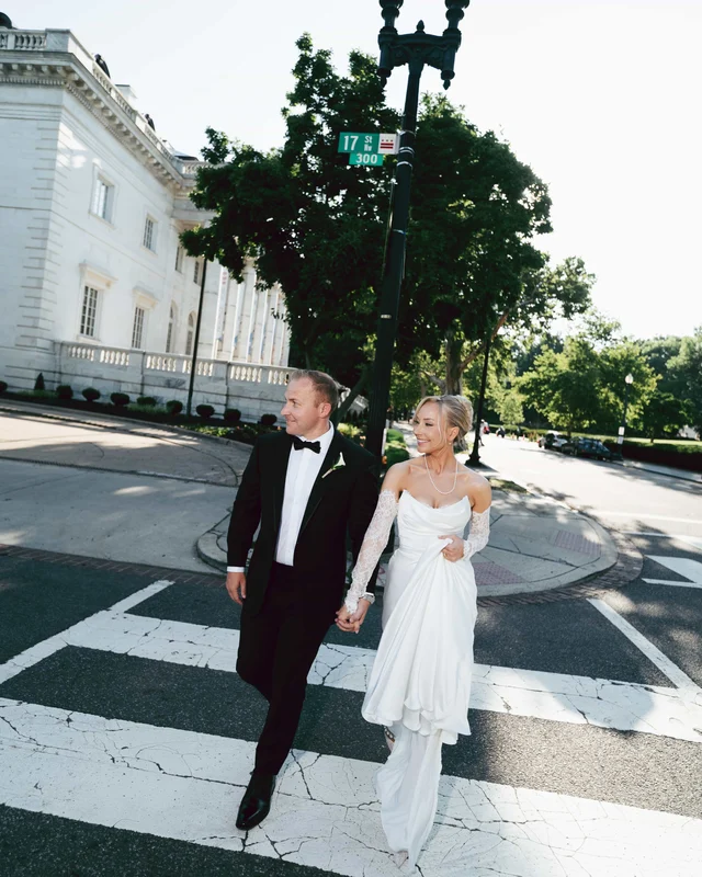 The newlyweds cross a sunlit crosswalk hand in hand beneath a street lamp, a marble facade rising behind the trees