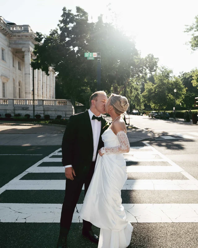 A kiss in the middle of the crosswalk, afternoon sun flaring through the trees behind them