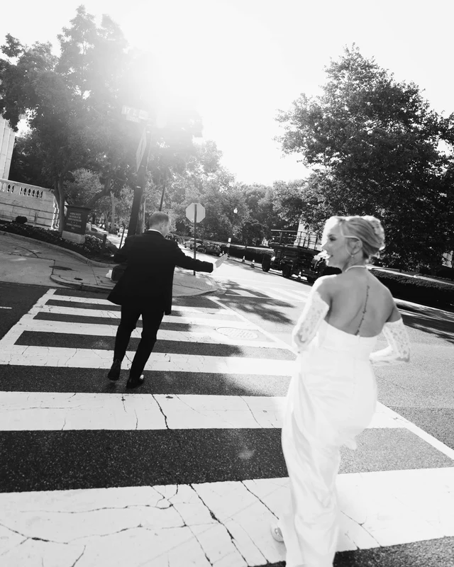 Black and white frame of the bride glancing back mid-crosswalk while the groom strides ahead, sun flooding the street