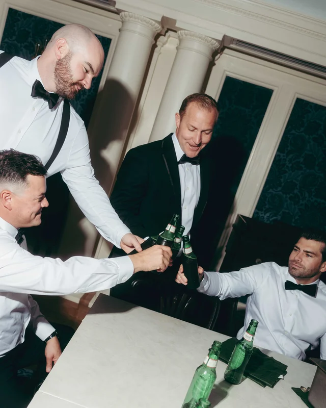 Four groomsmen clink green bottles over a table in a parlor lined with dark damask wallpaper
