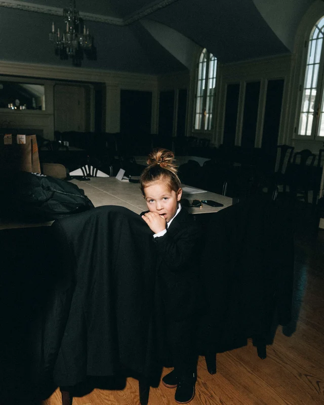 A small boy in a black suit and topknot waits beside draped garment bags in the dim reception hall