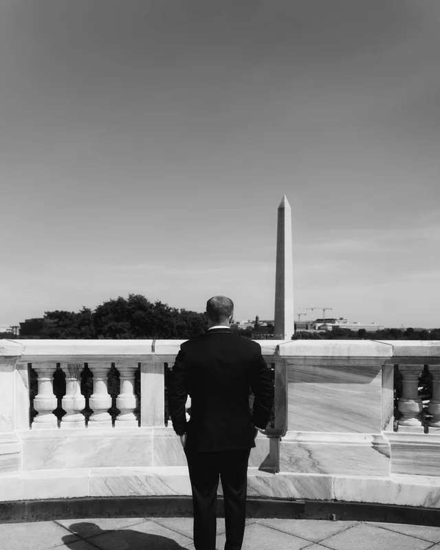 The groom stands alone at the rooftop balustrade in black and white, facing the distant obelisk