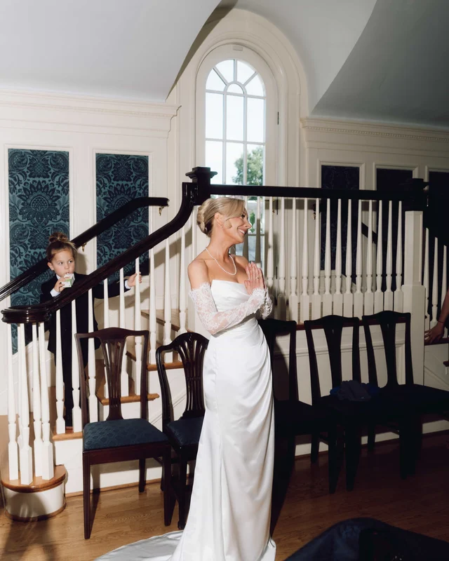 The bride presses her hands together and smiles across the staircase hall while a little boy peeks through the balusters