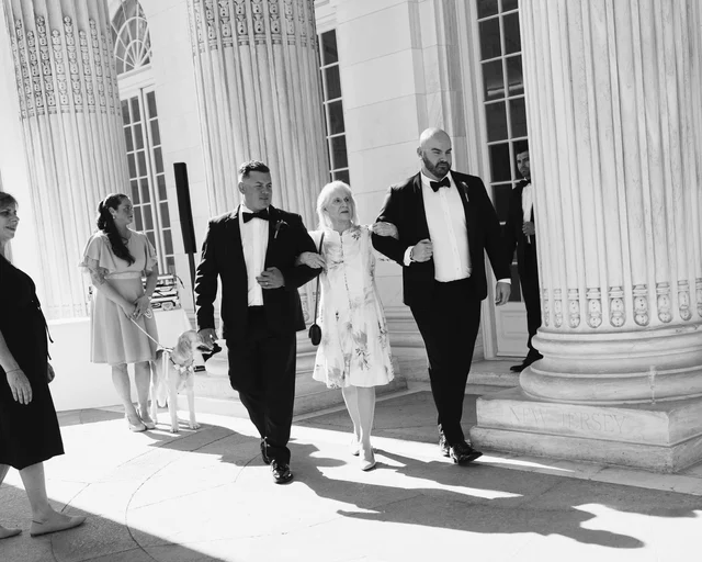 A tuxedoed groomsman escorts a woman in a floral dress past the colonnade, a leashed dog waiting at the edge of the frame
