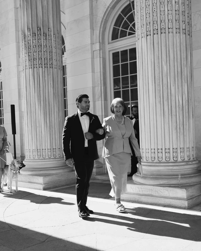 A dark-haired groomsman walks arm in arm with an older woman in a skirt suit beneath the towering fluted columns