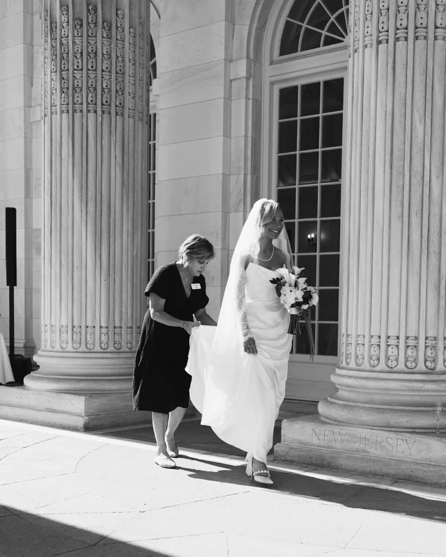 An attendant lifts the bride's train as she crosses the portico with her calla lily bouquet