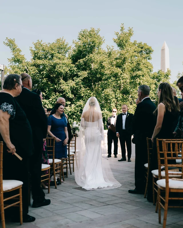 The bride starts down the aisle under a cathedral veil, guests turning as the groom waits beneath the trees