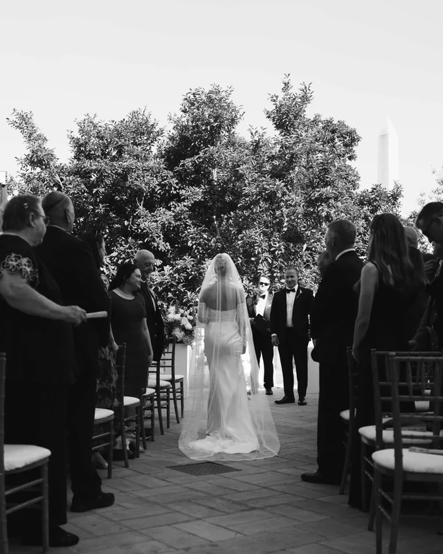 Black and white view down the aisle, the bride's veil trailing between rows of standing guests