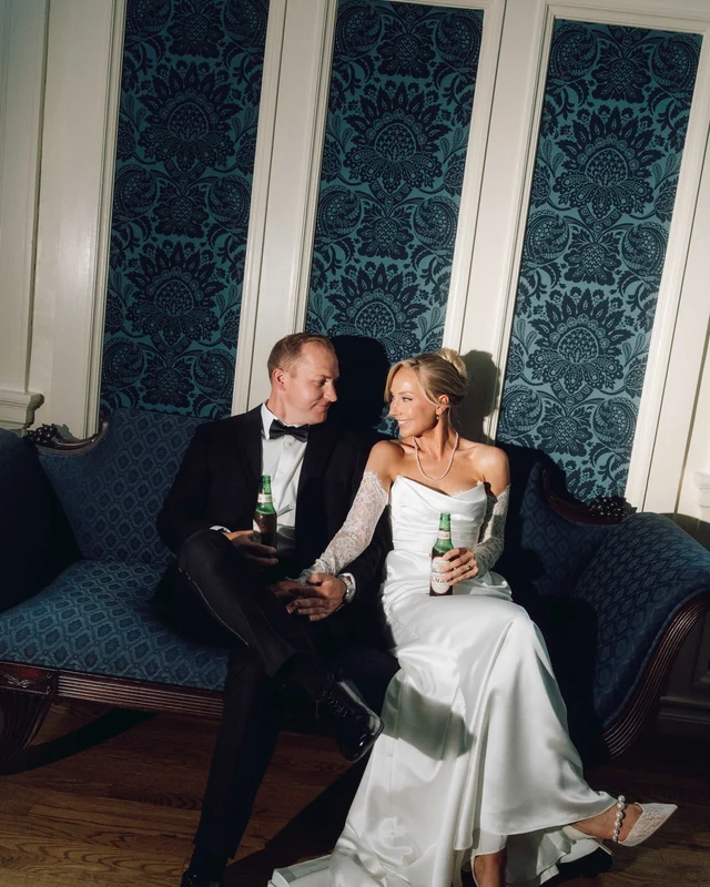 The newlyweds sink into a blue damask settee with cold beers, trading a look