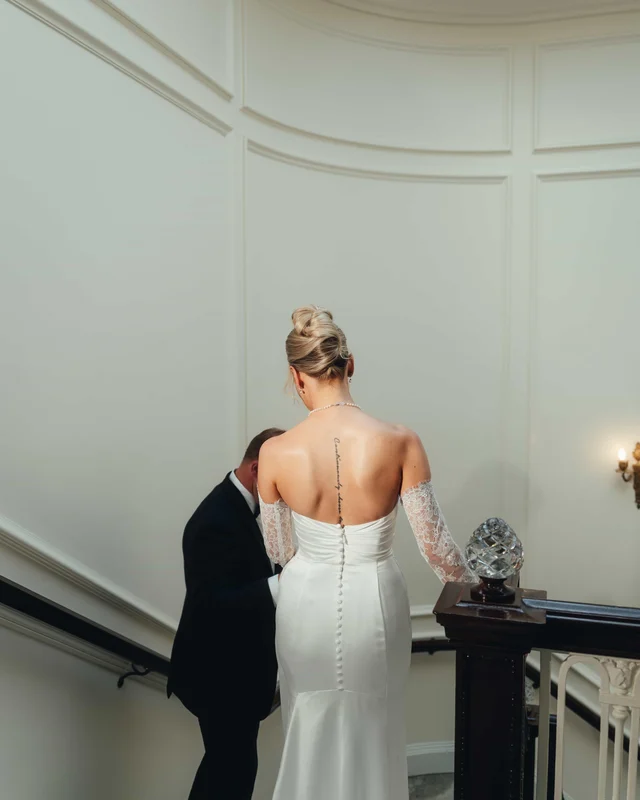 The bride from behind at the top of the stairs, buttoned satin and a script tattoo tracing her spine