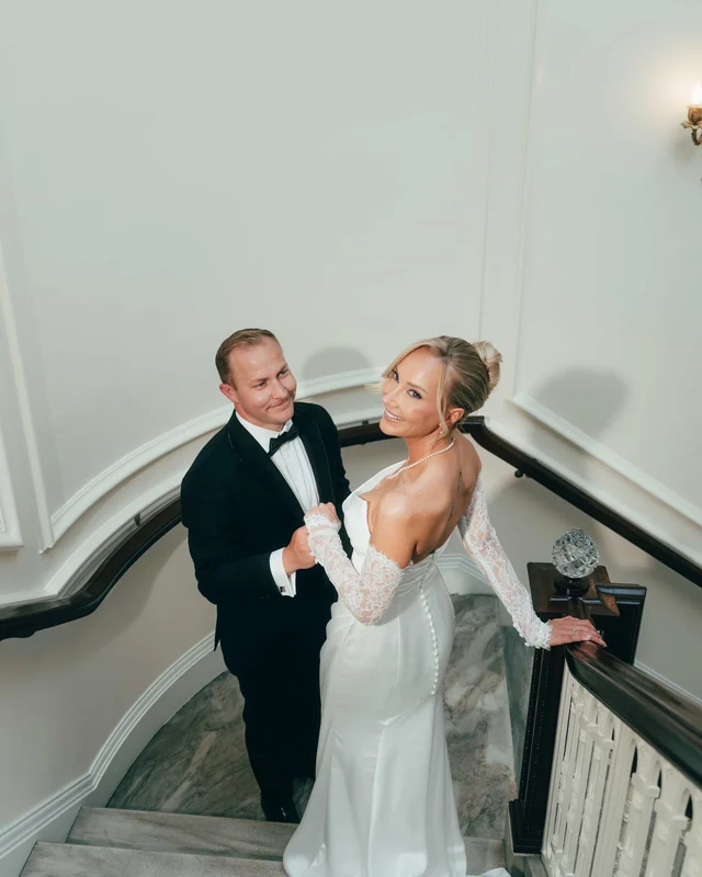Seen from above, the groom clasps the bride's hand on the staircase landing beside a crystal newel globe