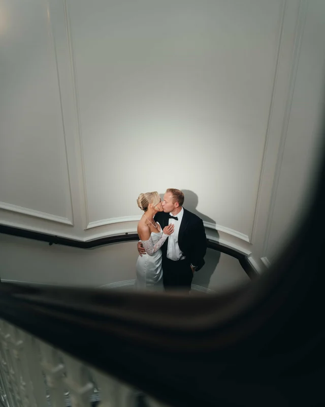 A quiet kiss on the stair landing, framed from above past the dark curve of the banister