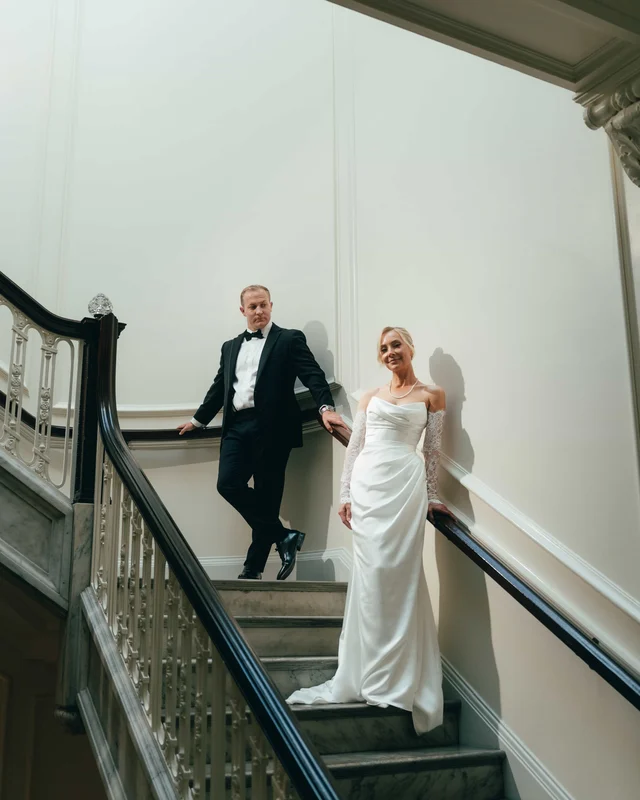The groom leans on both rails of the marble staircase above the bride, who pauses on the steps below