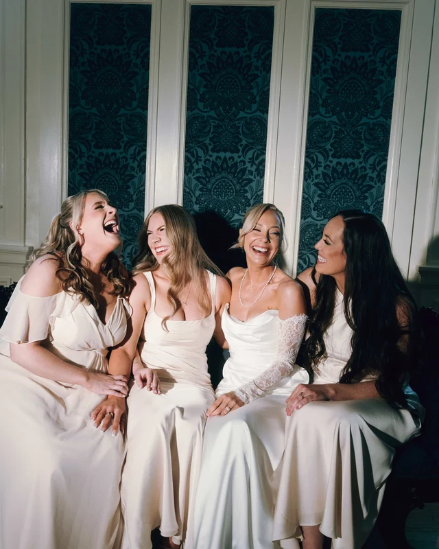 The bride and three bridesmaids collapse into laughter together on a settee against teal damask panels