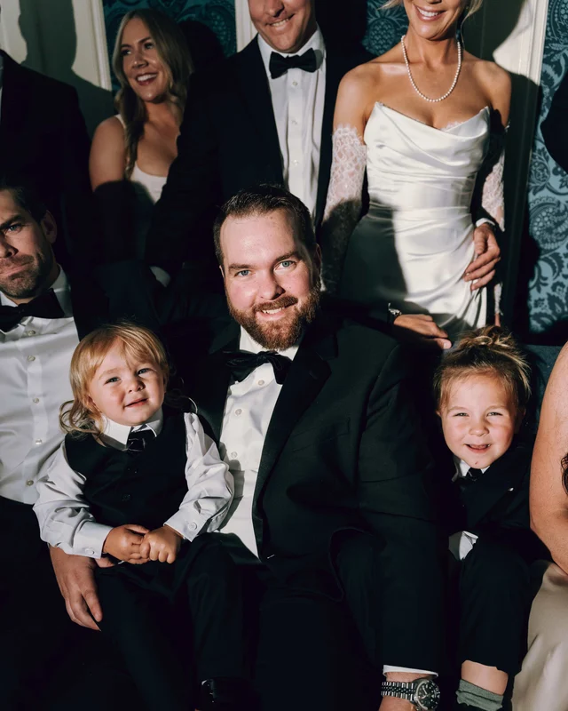 A bearded groomsman holds two small children in miniature suits on his lap during wedding party portraits