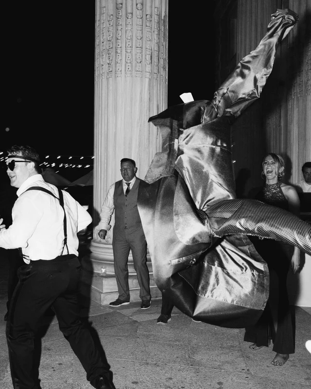 A suit jacket flies through the air mid-dance as a guest in suspenders lunges past the column, another laughing barefoot