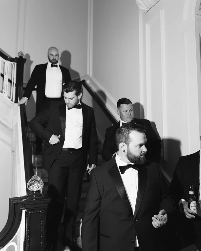 The groom and groomsmen file down the staircase in black and white, a beer bottle in hand at the rail