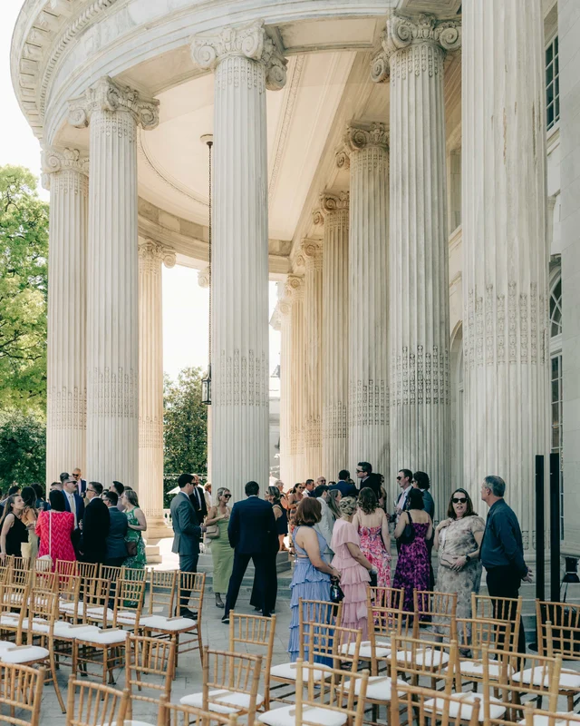 Guests in summer colors gather beneath the curved colonnade before the ceremony, gold chairs waiting in the foreground