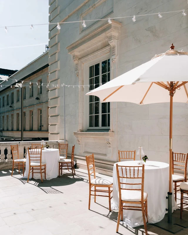 Cafe tables and a white umbrella on the sunlit terrace, string lights strung along the carved stone facade