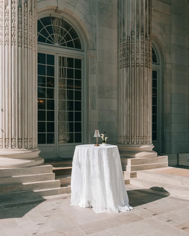 A single linen-draped cocktail table with a tiny lamp waits between colossal columns and arched glass doors