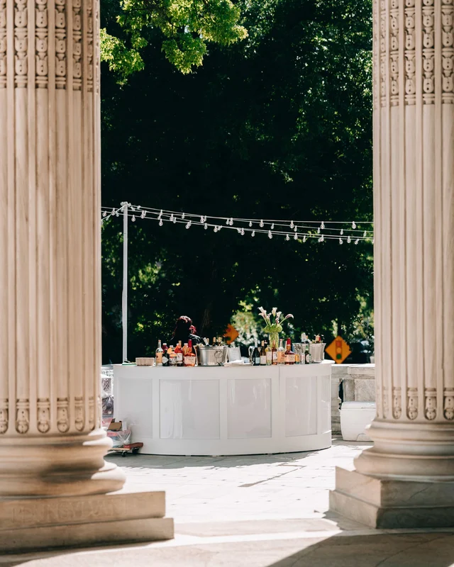 The white curved bar glimpsed between two enormous fluted column bases, string lights sagging toward the dark trees