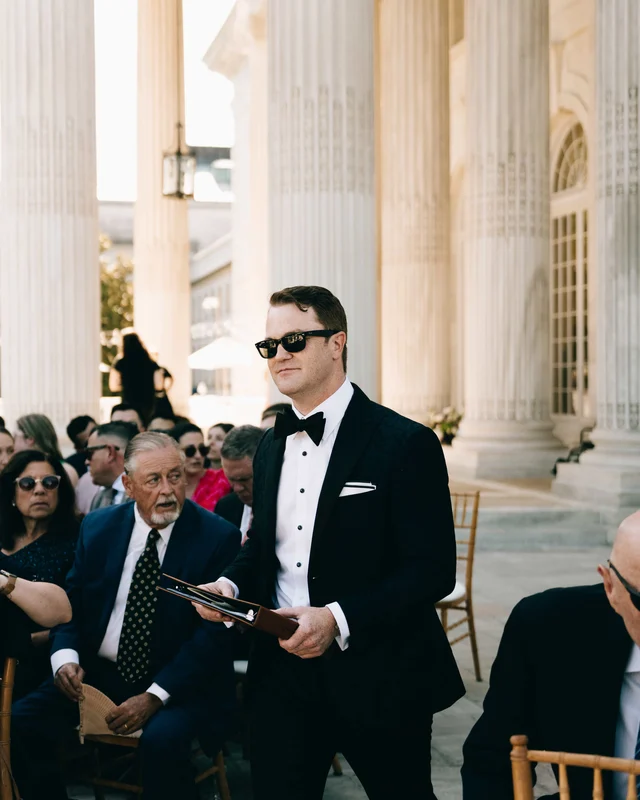 A man in sunglasses carries a leather folio down the aisle past seated guests before the ceremony
