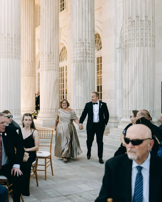 The groom walks in hand in hand with a woman in a champagne gown, guests rising along the aisle