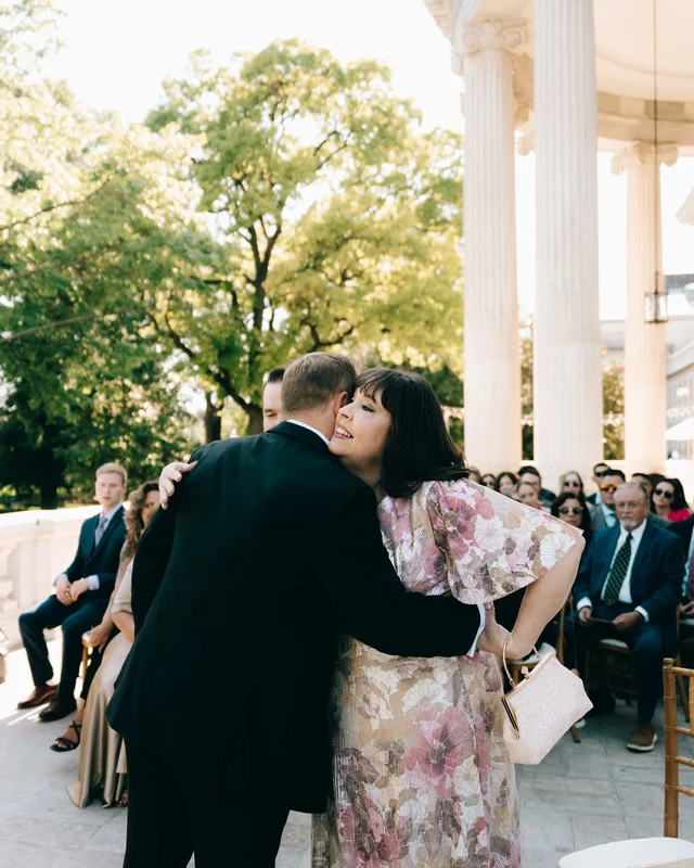 A tuxedoed man embraces a woman in a blush floral dress before taking his place, guests looking on