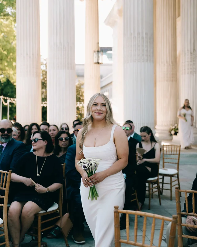 A bridesmaid in ivory carries calla lilies up the aisle past rows of seated guests