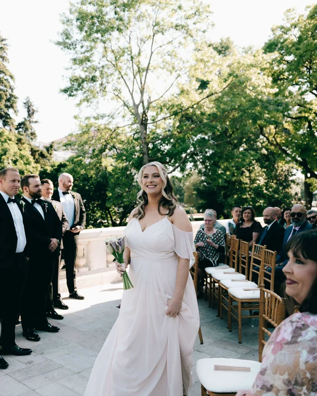 A bridesmaid in a blush off-shoulder gown processes past the waiting groomsmen, bouquet low at her hips