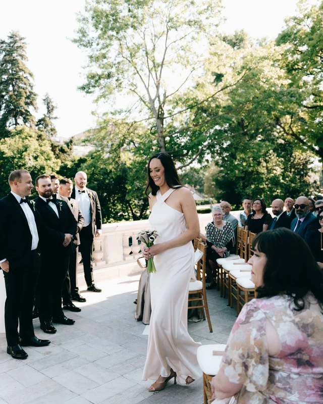 A bridesmaid in a champagne halter gown carries her calla lilies between the groomsmen and the seated rows