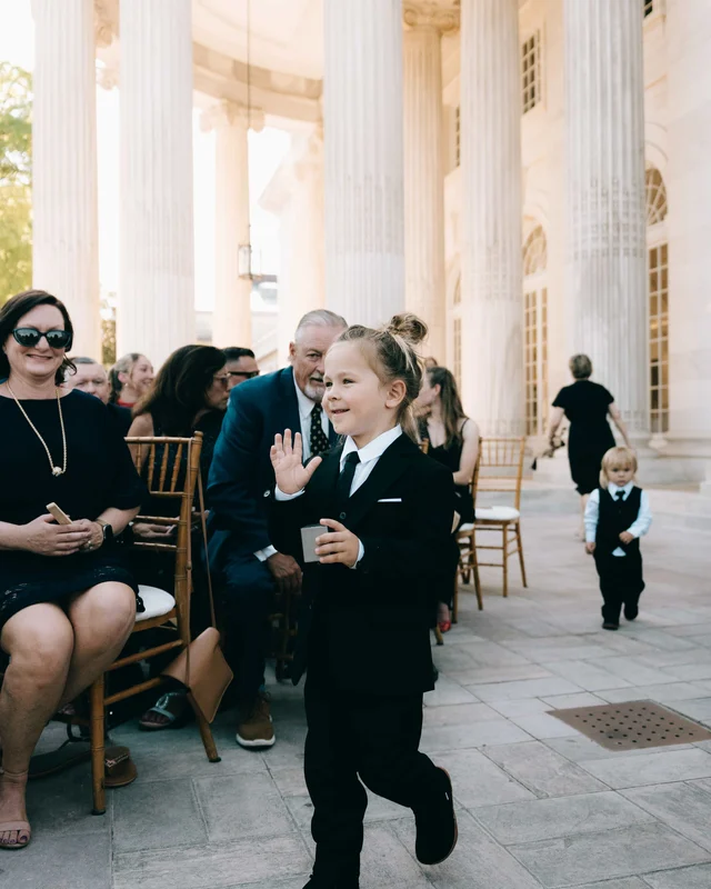The ring bearer waves to the crowd mid-aisle, a second small boy trailing far behind him
