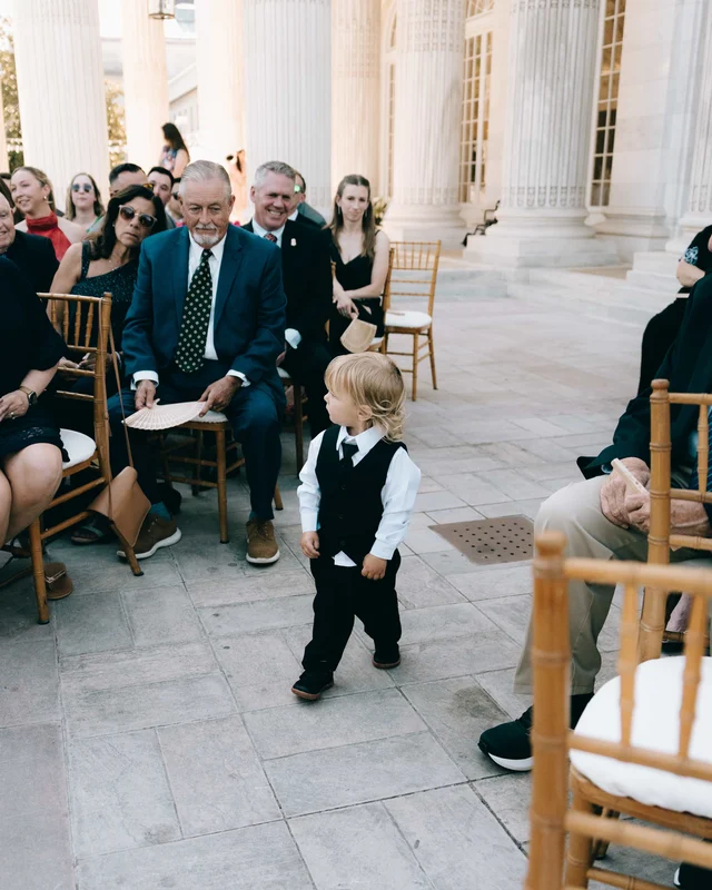 A toddler in a vest and tie wanders the aisle while seated guests grin and fan themselves