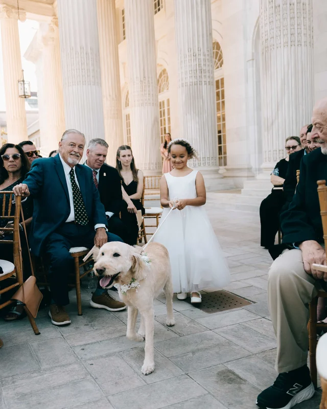 The flower girl in white leads the couple's dog up the aisle on a ribbon leash