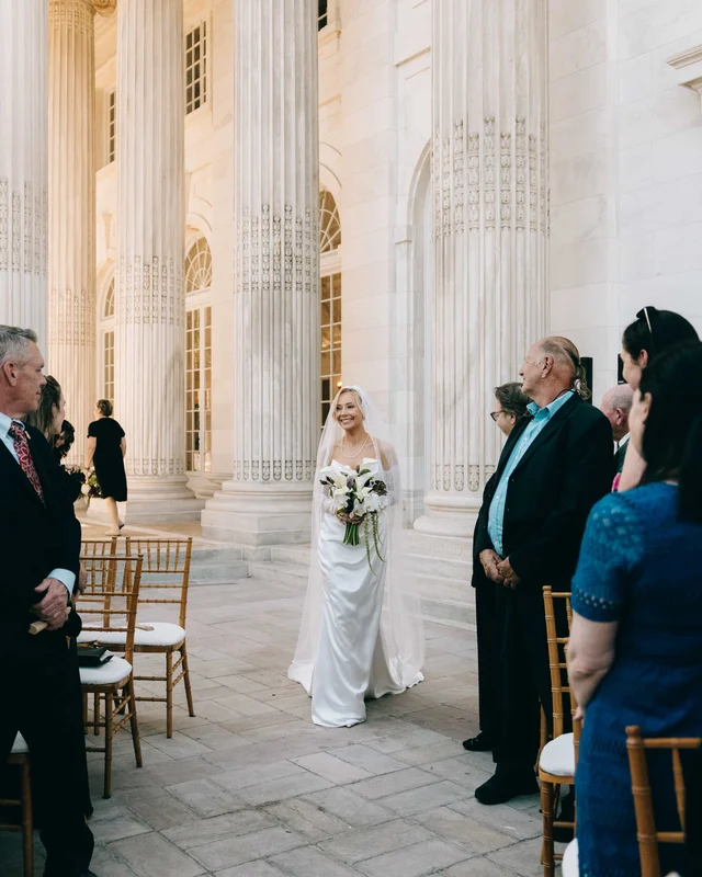 The bride emerges alone from the towering colonnade, guests rising at the edge of the aisle