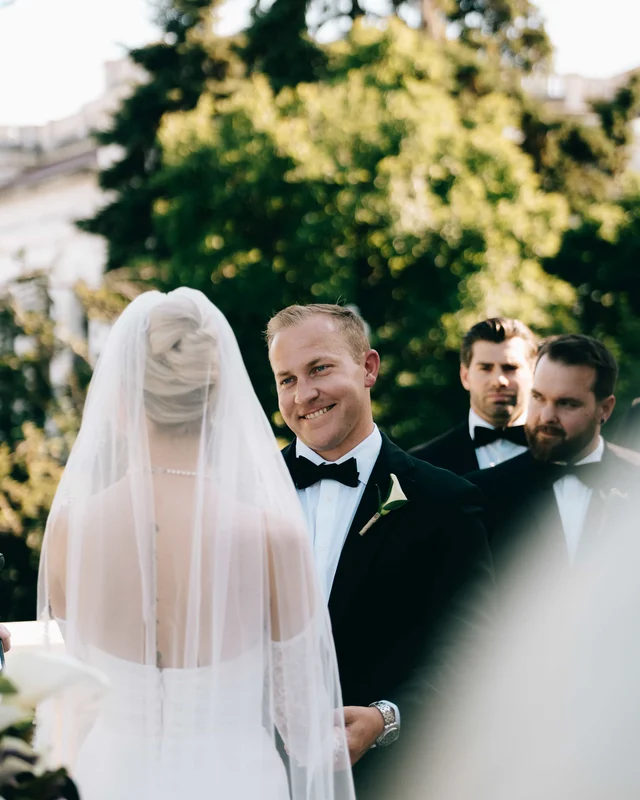 Over the bride's veiled shoulder, the groom grins at her from the head of the aisle