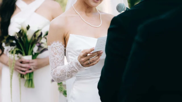 The bride's lace-sleeved hands hold a vow card at the microphone, her ring catching the light