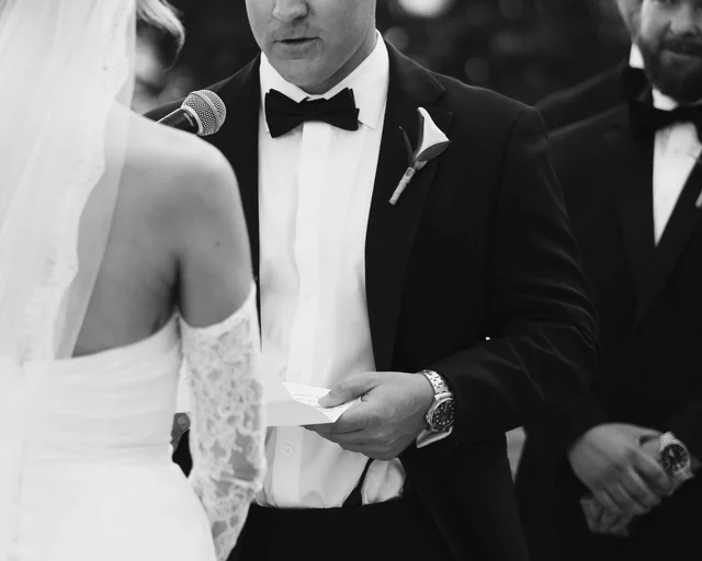 Close black and white frame of the groom reading his vows from a card at the microphone