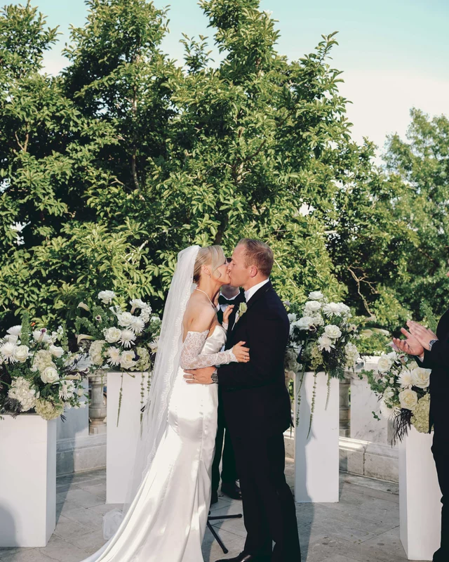 The first kiss at the altar between pedestal arrangements of white flowers, trees rising behind