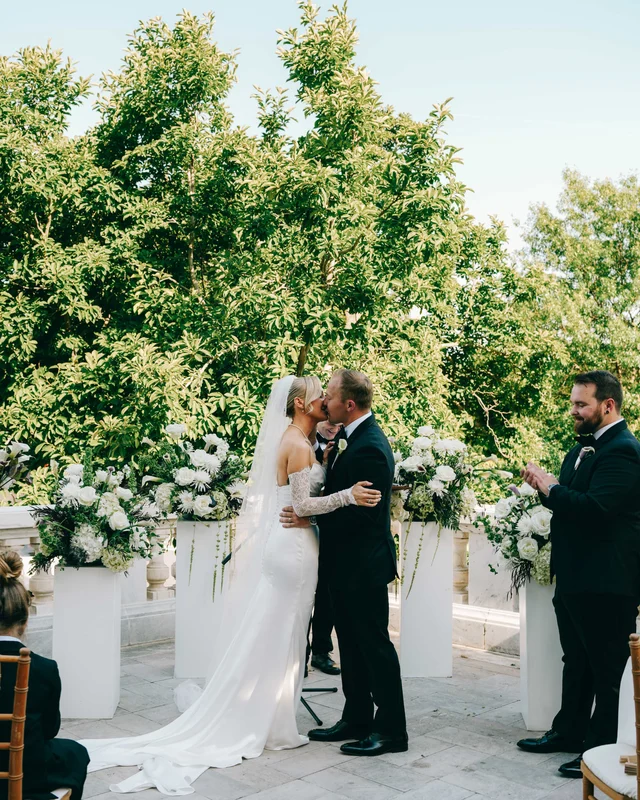 The couple holds their first kiss beside the flower-topped pedestals as applause breaks out at the edge of the frame