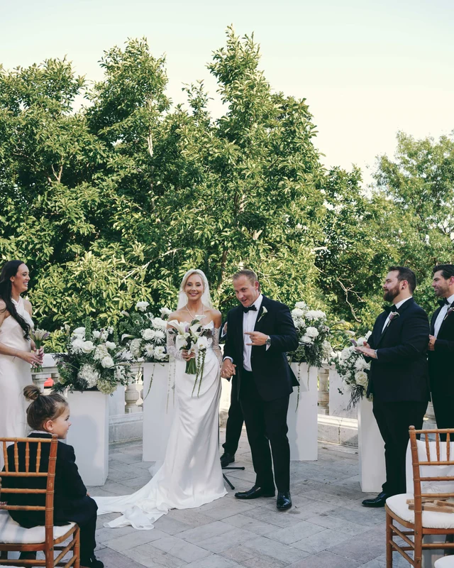 Just married, the groom points at his laughing bride while a small boy watches from a chair