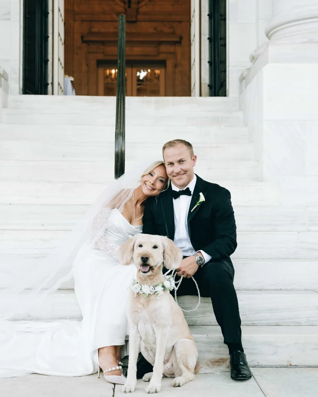 The couple sits on the marble steps with their dog, the bride's head resting on the groom's shoulder