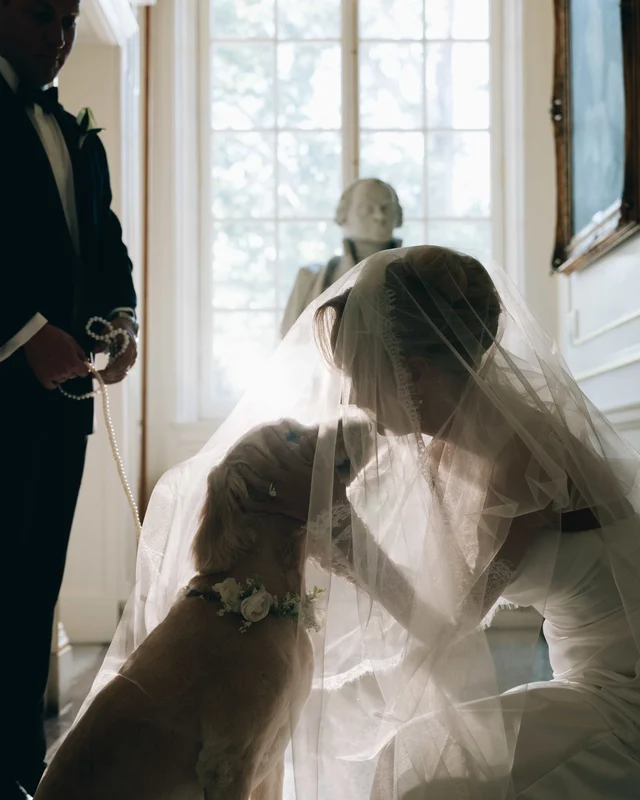 Beneath her veil, the bride bends to kiss the dog's head while the groom holds the pearl leash