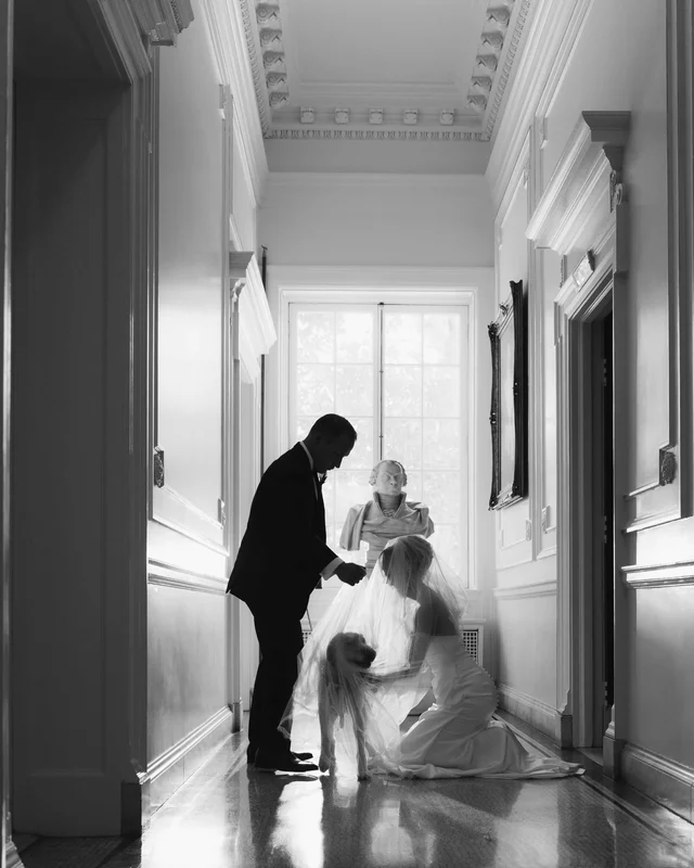 Black and white corridor scene, the bride kneeling with the dog under her veil beside a marble bust