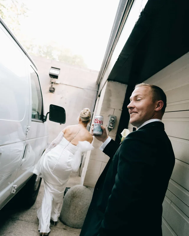The groom raises a silver beer can to the camera as the bride heads up the alley