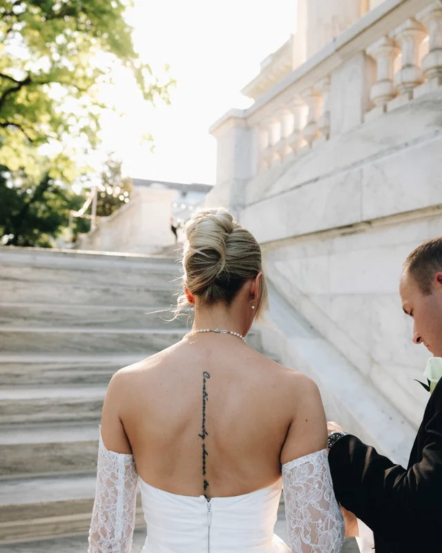 The bride's French twist and spine tattoo from behind as the groom fastens her gown on the steps