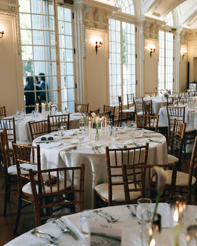Round tables dressed in white linen with calla lilies and taper candles beneath tall arched windows